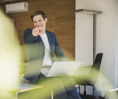 Glücklicher Geschäftsmann mit Laptop im Büro, der mit dem Finger zeigt - UUF25780