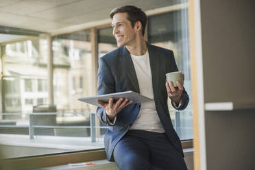 Confident businessman holding tablet and coffee mug at the window in office - UUF25798