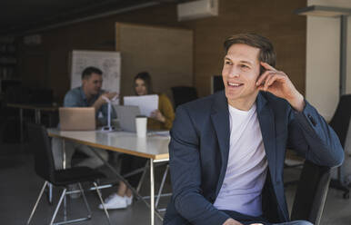 Confident businessman sitting on chair in office with colleagues in background - UUF25801