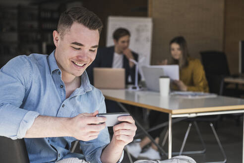 Lächelnder Geschäftsmann, der im Büro sein Smartphone benutzt, mit Kollegen im Hintergrund - UUF25803