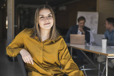 Selbstbewusste Geschäftsfrau, die auf einem Stuhl im Büro sitzt, mit Kollegen im Hintergrund - UUF25804