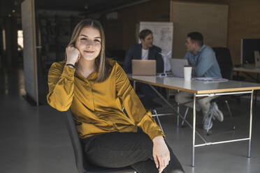 Selbstbewusste Geschäftsfrau, die auf einem Stuhl im Büro sitzt, mit Kollegen im Hintergrund - UUF25805