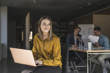 Businesswoman using laptop in office with colleagues in background - UUF25806