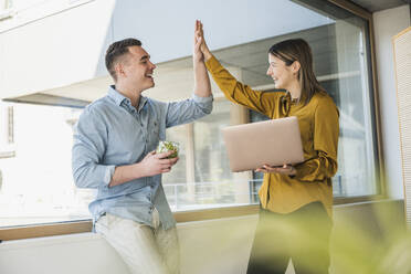 Businessman and businesswoman with laptop high fiving at the window in office - UUF25832