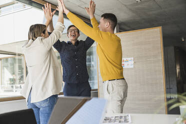 Glückliche Geschäftsleute, die mit erhobenen Armen im Büro zusammenstehen - UUF25854