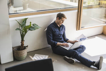 Businessman sitting on the floor in office using laptop - UUF25861