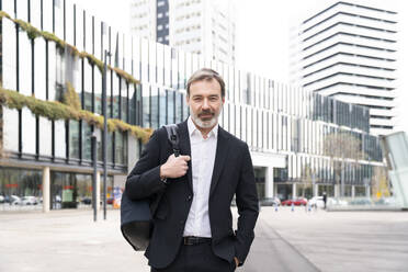 Lächelnder Geschäftsmann mit Hand in der Tasche vor einem modernen Bürogebäude stehend - OIPF01505