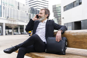 Smiling businessman talking on smart phone sitting with laptop bag on bench - OIPF01512