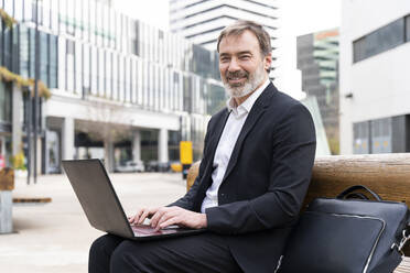 Lächelnder Geschäftsmann mit Laptop auf einer Bank sitzend - OIPF01514