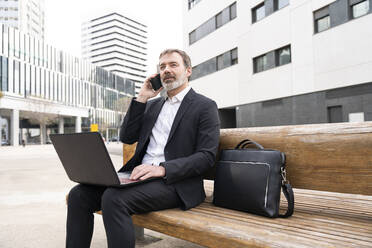 Älterer Geschäftsmann, der mit einem Laptop auf einer Bank sitzt und mit einem Mobiltelefon spricht - OIPF01515