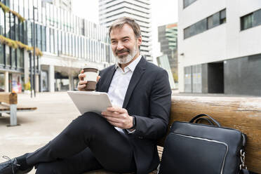 Lächelnder reifer Geschäftsmann mit Tablet-PC und Einweg-Kaffeebecher auf einer Bank sitzend - OIPF01519