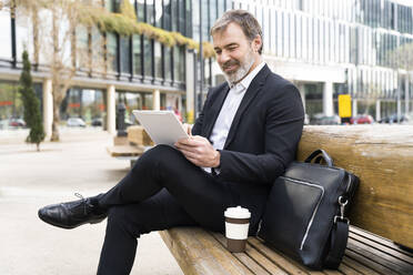 Smiling mature businessman using tablet PC sitting on bench - OIPF01521