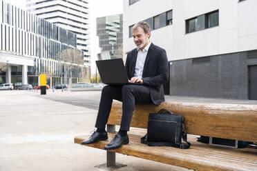 Smiling businessman using laptop sitting on bench in front of office building - OIPF01526