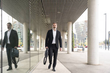 Mature businessman carrying laptop walking by glass wall - OIPF01528