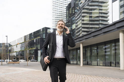 Lächelnder reifer Geschäftsmann mit Laptoptasche, der mit seinem Smartphone telefoniert und vor einem Bürogebäude spazieren geht - OIPF01530