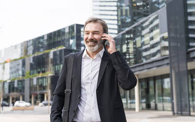 Glücklicher Geschäftsmann, der vor einem Bürogebäude mit einem Mobiltelefon spricht - OIPF01531