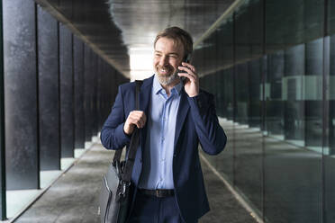 Happy businessman with laptop bag talking on smart phone standing in corridor - OIPF01535