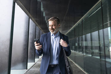 Lächelnder reifer Geschäftsmann, der im Korridor stehend mit seinem Mobiltelefon im Internet surft - OIPF01538