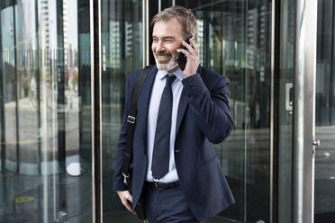 Smiling mature businessman talking on smart phone walking out from revolving door - OIPF01563