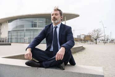 Businessman meditating sitting crossed-legged on wall - OIPF01571