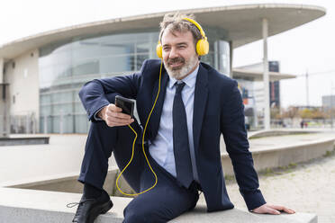 Entspannter Geschäftsmann, der mit Kopfhörern an der Wand sitzend Musik hört - OIPF01574