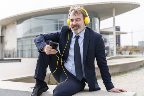 Entspannter Geschäftsmann, der mit Kopfhörern an der Wand sitzend Musik hört - OIPF01574