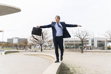 Businessman walking on wall holding laptop bag at office park - OIPF01581