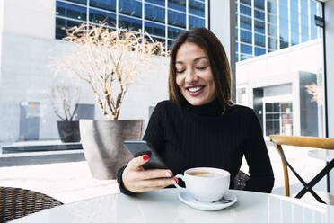 Smiling businesswoman with coffee cup using smart phone sitting in cafe - PNAF03739