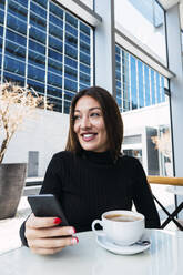 Happy businesswoman with smartphone and coffee cup sitting at table at cafe - PNAF03740