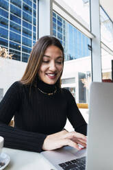 Smiling young businesswoman using laptop sitting in cafe - PNAF03743