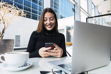 Smiling young businesswoman using smart phone sitting with laptop in cafe - PNAF03744