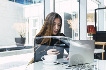 Geschäftsfrau sitzt mit Laptop und Smartphone durch ein Glasfenster in einem Café gesehen - PNAF03753