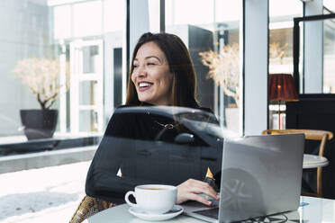 Happy businesswoman with laptop seen through glass window sitting in cafe - PNAF03754