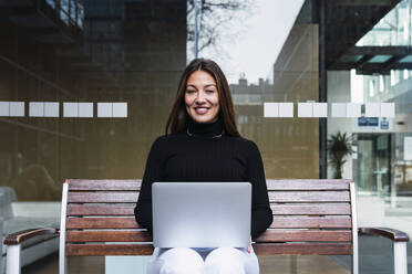 Glückliche junge Geschäftsfrau mit Laptop auf einer Bank vor einer Glaswand sitzend - PNAF03764