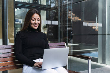 Smiling businesswoman using laptop sitting on bench - PNAF03765