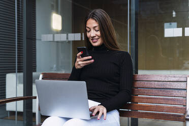 Lächelnde junge Geschäftsfrau, die ein Smartphone benutzt und mit einem Laptop auf einer Bank sitzt - PNAF03766