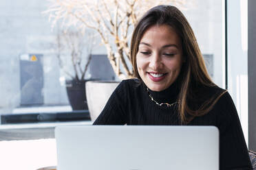 Lächelnde junge Geschäftsfrau mit braunem Haar, die in einem Café einen Laptop benutzt - PNAF03779