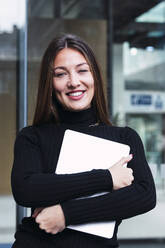 Smiling young businesswoman with laptop standing in front of glass wall - PNAF03784