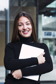Lächelnde junge Geschäftsfrau mit Laptop vor einer Glaswand stehend - PNAF03784