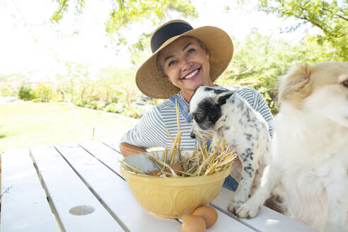 Smiling woman and dogs sitting with eggs bowl at table in garden - ESTF00011