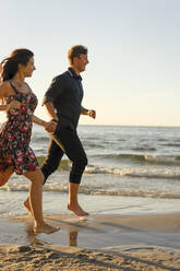Glückliches Paar hält sich an den Händen und läuft an einem sonnigen Tag am Strand - SSGF00766
