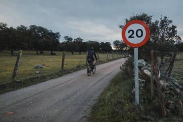 Radfahrer fährt bei Sonnenaufgang auf der Straße an einem Schild mit Geschwindigkeitsbegrenzung vorbei - DMGF00696