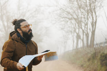 Mature bearded man wearing eyeglasses standing with map in forest - MRRF02005