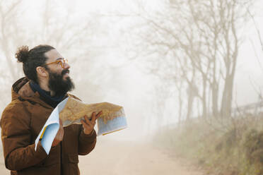 Happy bearded man with map standing in forest - MRRF02006