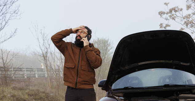 Besorgter Mann, der am Auto stehend mit seinem Handy spricht - MRRF02007