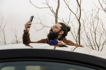 Mature bearded man checking smart phone standing behind car - MRRF02009