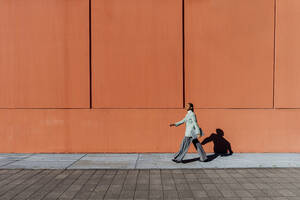 Junge Geschäftsfrau mit Laptop geht an einer orangefarbenen Wand vorbei - MEUF05176