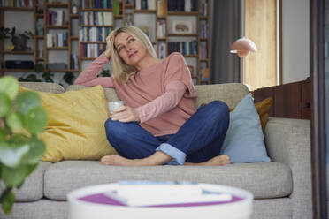 Smiling woman with coffee cup sitting cross-legged on sofa at home - RBF08876