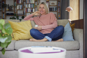 Happy woman with coffee cup sitting on sofa at home - RBF08877