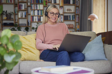 Glückliche Frau mit Headset und Laptop auf dem Sofa zu Hause - RBF08879
